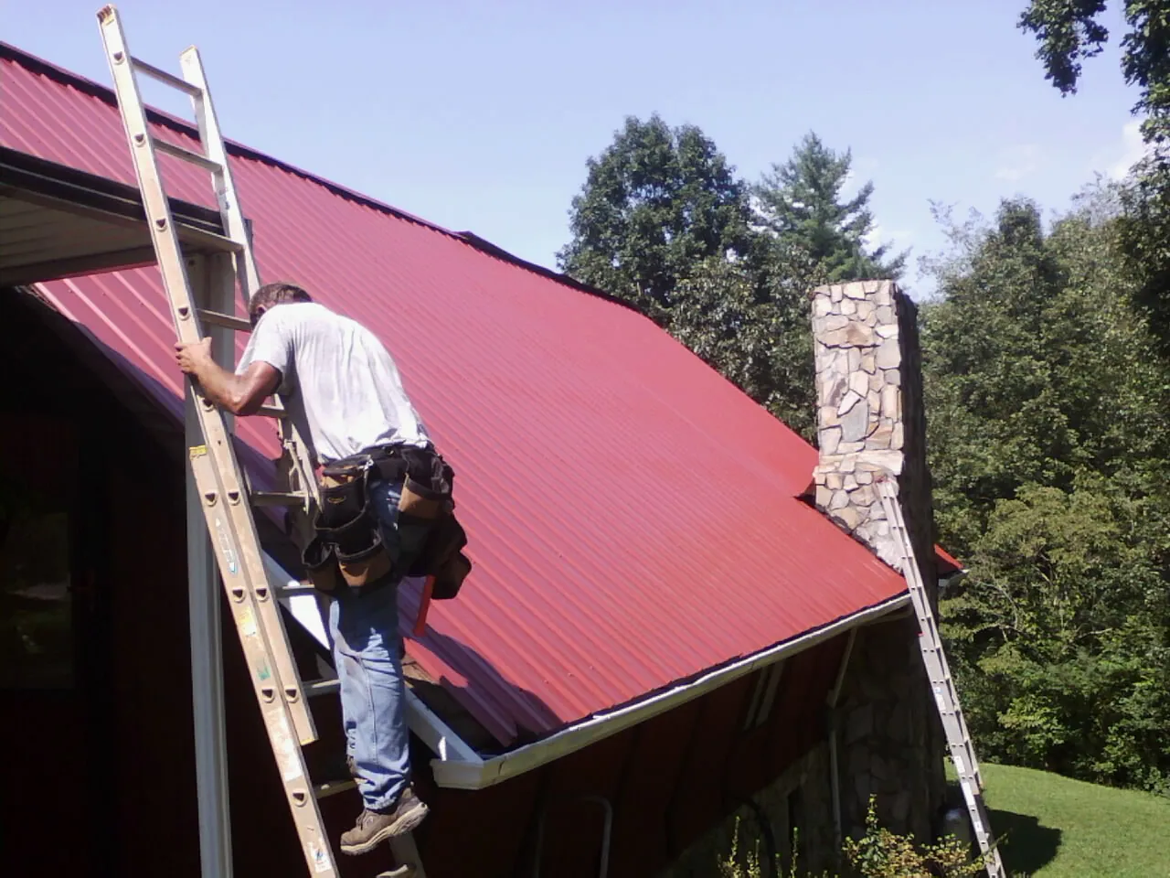 Roofer on a ladder beside a red metal roof and stone chimney — Soffit & Fascia Repair by Roofing Brothers in Birdsboro